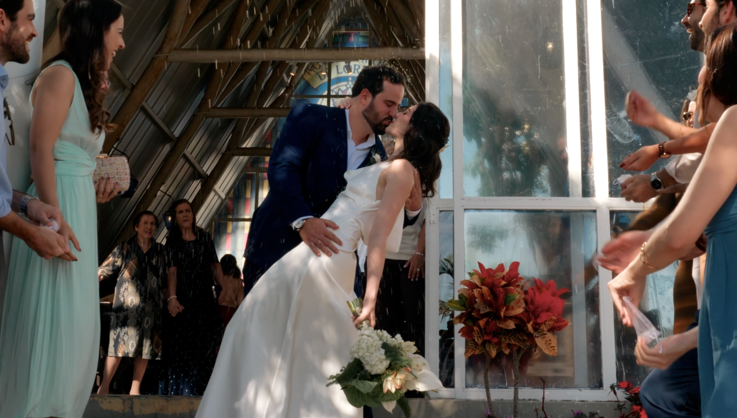Bride and groom share first kiss at rustic barn wedding surrounded by cheering wedding party.