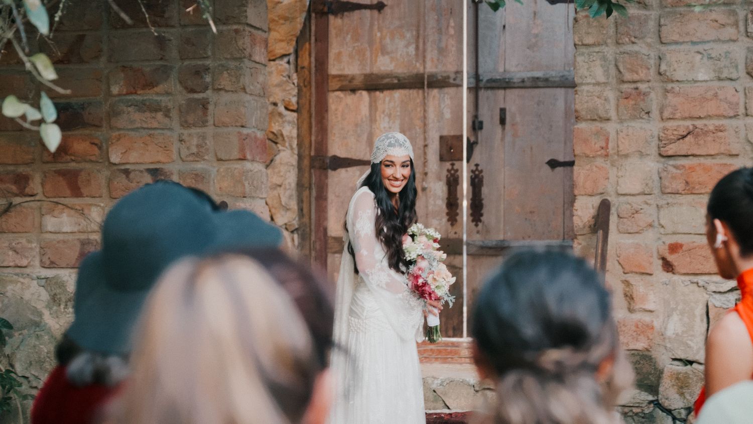 Smiling bride in white dress and gray beanie holds bouquet at rustic outdoor wedding ceremony.
