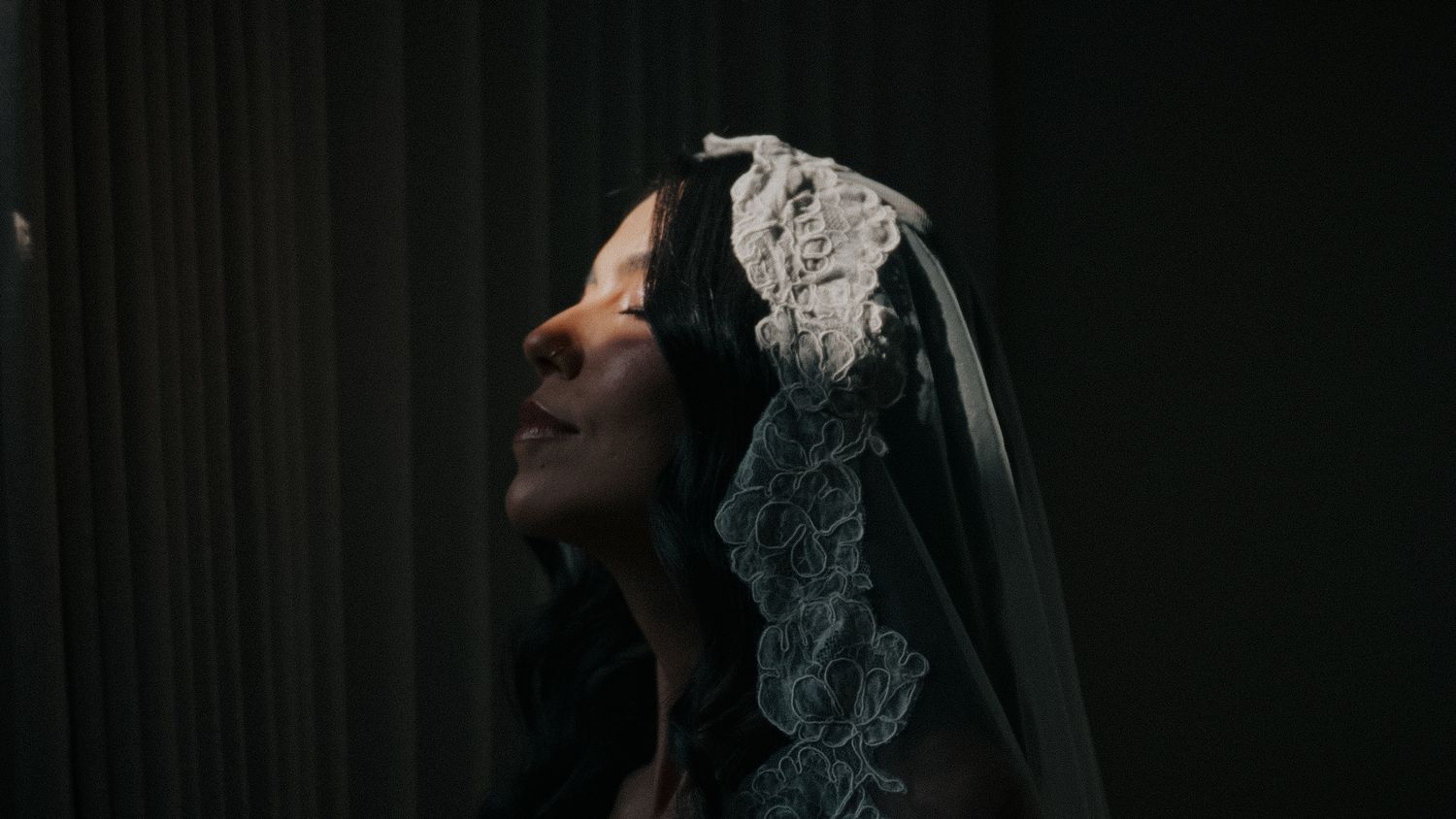 A bride wearing an elegant lace veil looks upward in a dimly lit room, her profile illuminated softly.