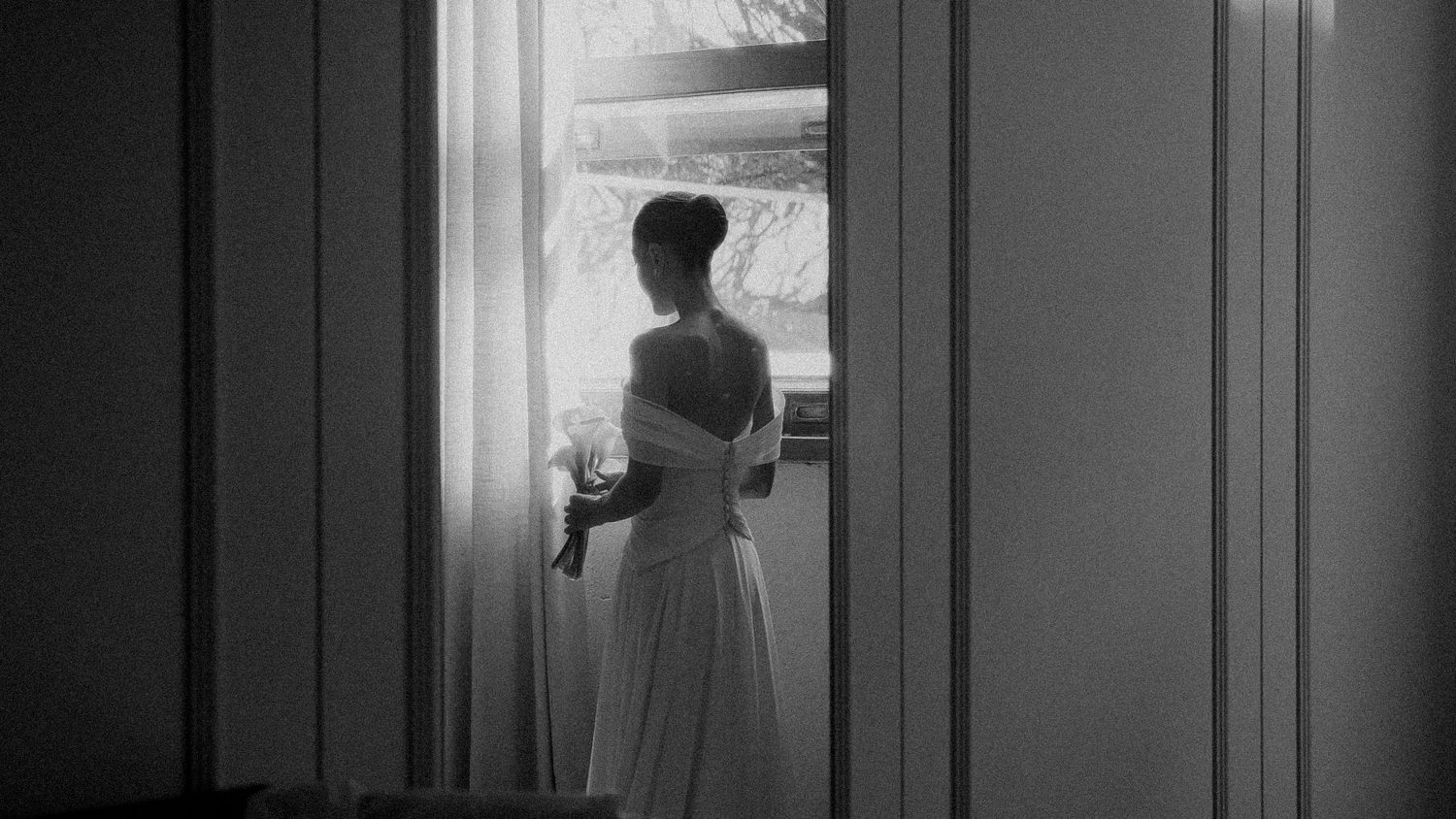 Black and white photo of a woman in a white off-shoulder gown standing by a bright window holding flowers.