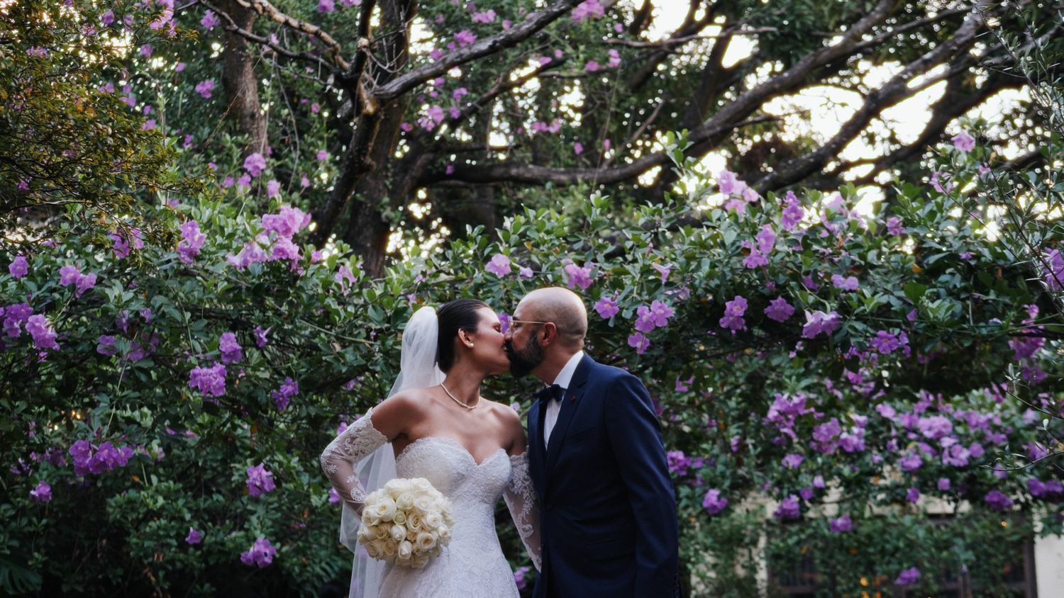 Bride and groom kissing under blooming purple flower tree in romantic outdoor wedding portrait.