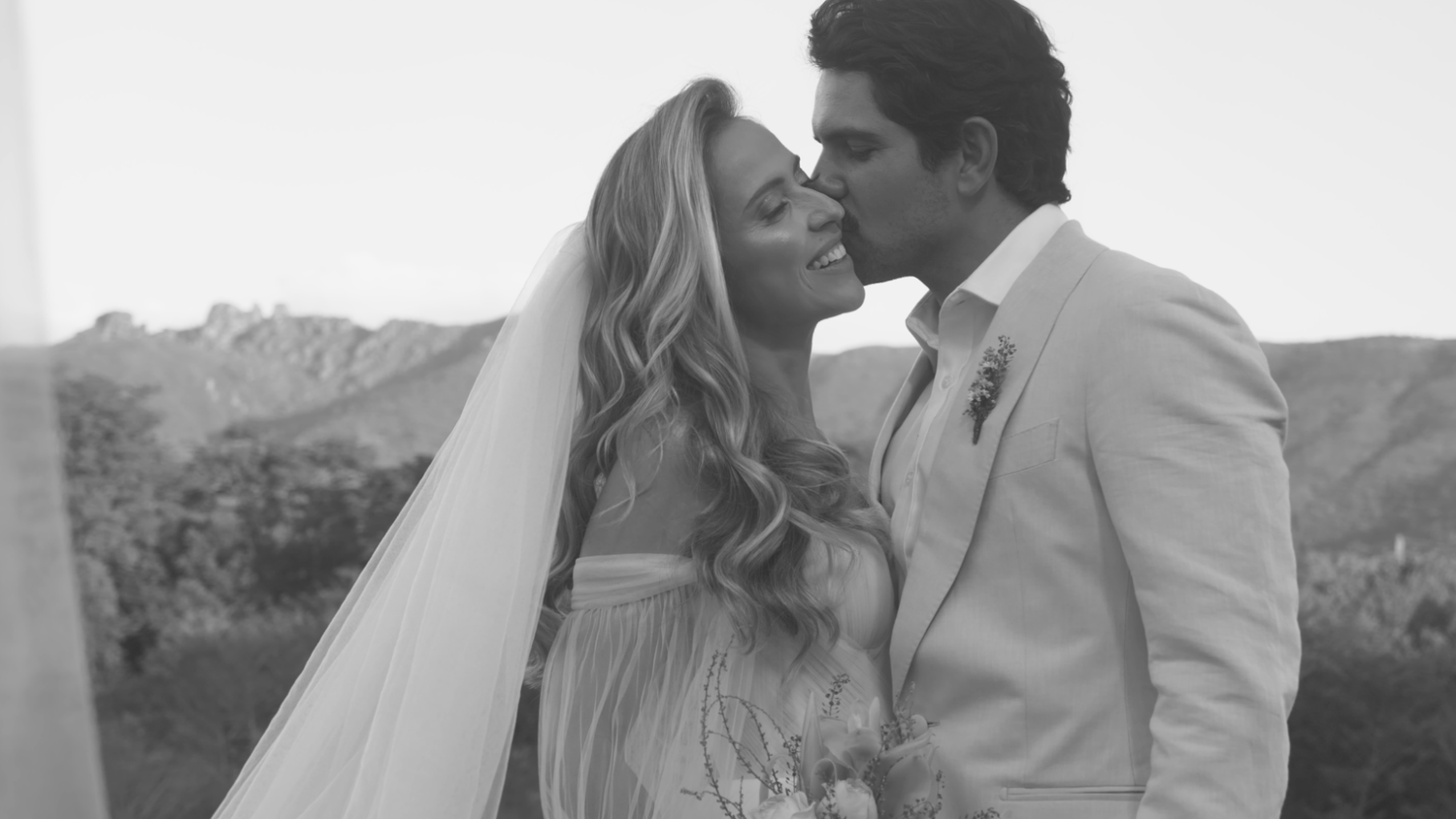 Bride and groom share romantic kiss with mountain backdrop in black and white wedding portrait.