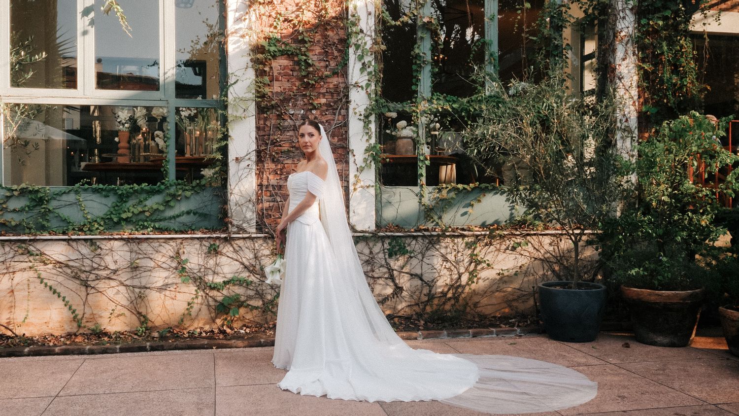 Bride in elegant white off-shoulder wedding gown with long veil standing in a lush garden courtyard.