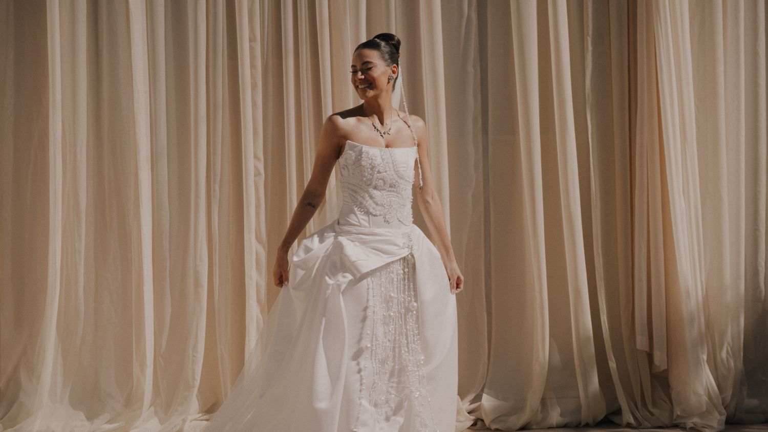 Bride in elegant white wedding gown with veil, posing gracefully in front of flowing beige curtains.