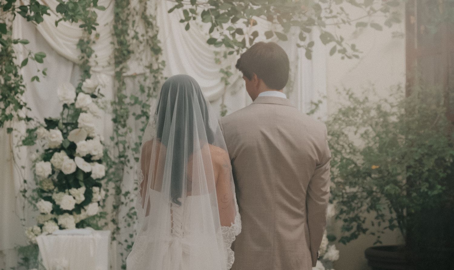 A bride and groom stand before a floral wedding altar adorned with white roses and greenery.