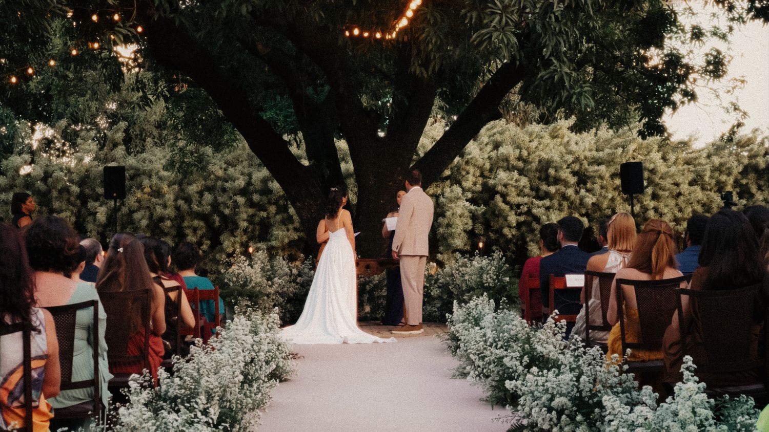Bride walks down white floral aisle toward groom under large tree with string lights at dusk.