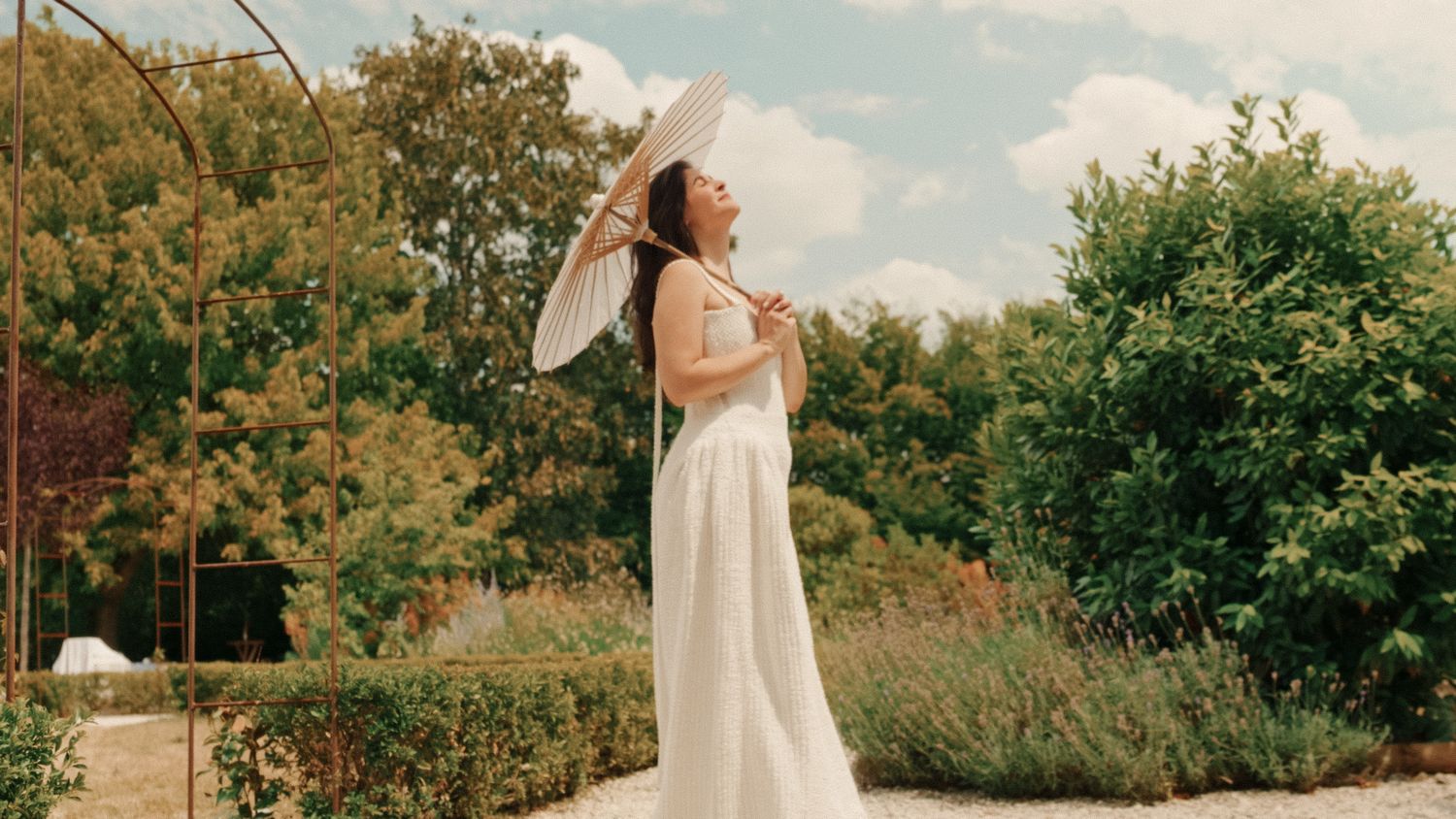 Woman in white dress holding parasol in lush garden with cloudy sky backdrop.