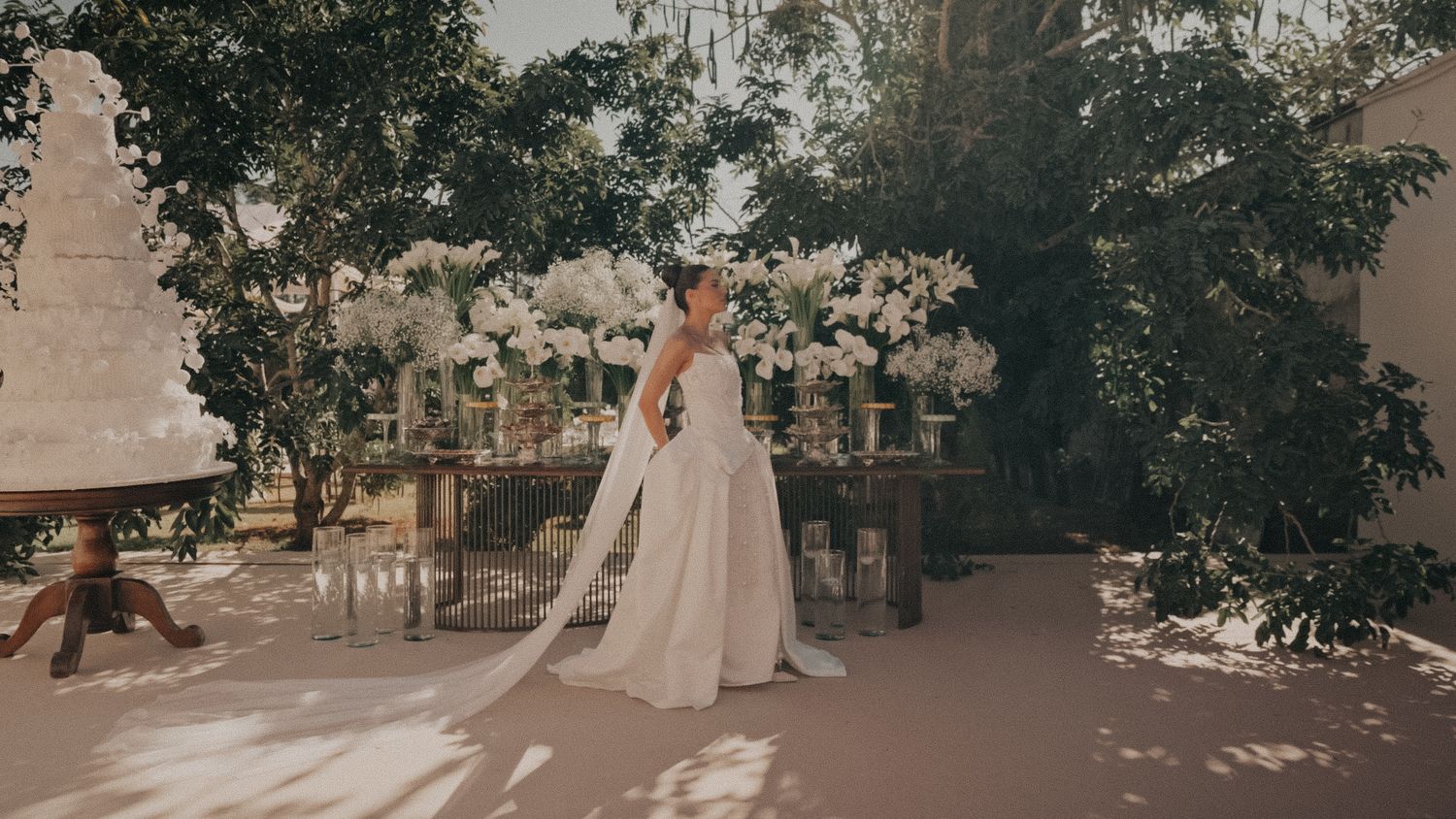 Bride in elegant white gown with flowing veil stands among lush trees and white floral wedding decorations.