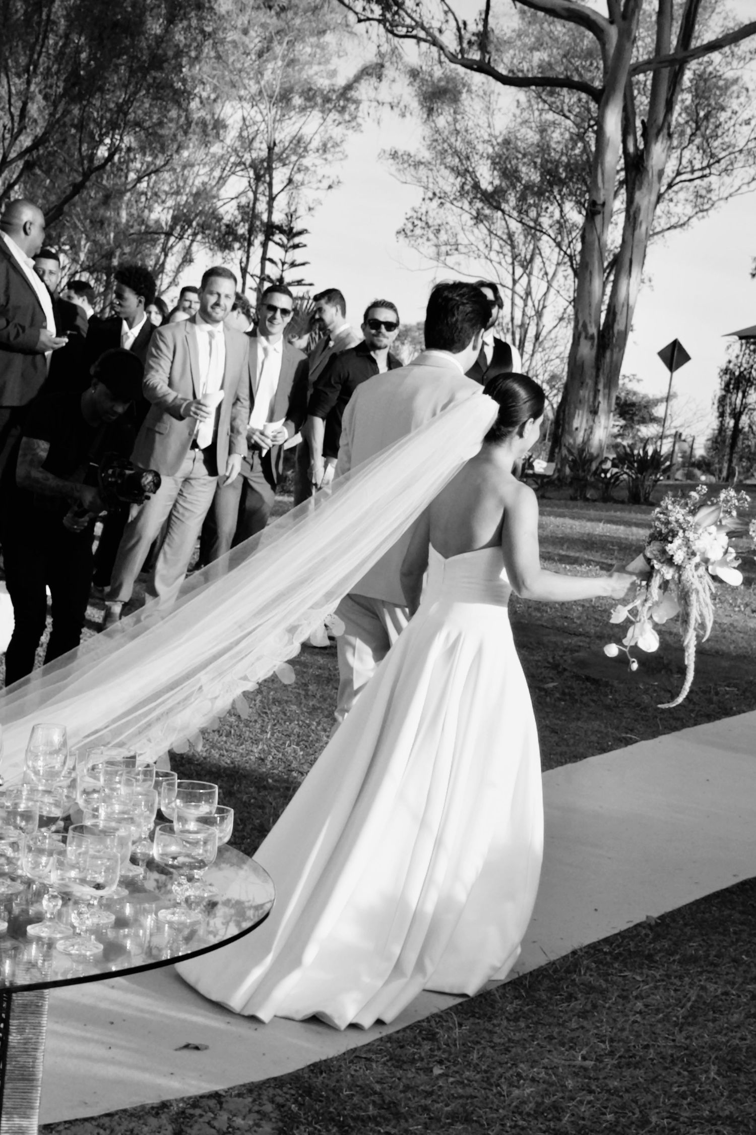Black and white photo of bride with long flowing veil walking toward guests at outdoor ceremony.
