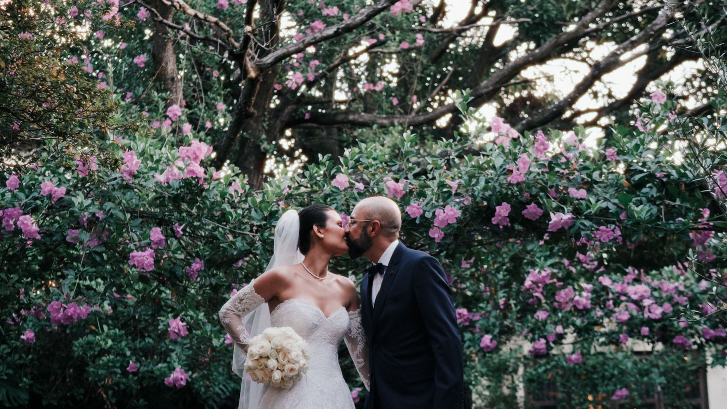 Bride and groom share a romantic kiss surrounded by lush pink flowering trees in a garden wedding setting.
