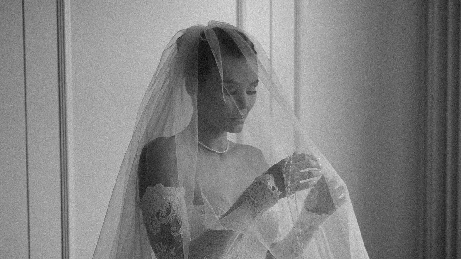 Black and white photo of a bride in a lace gown and veil, looking down gracefully near a window.