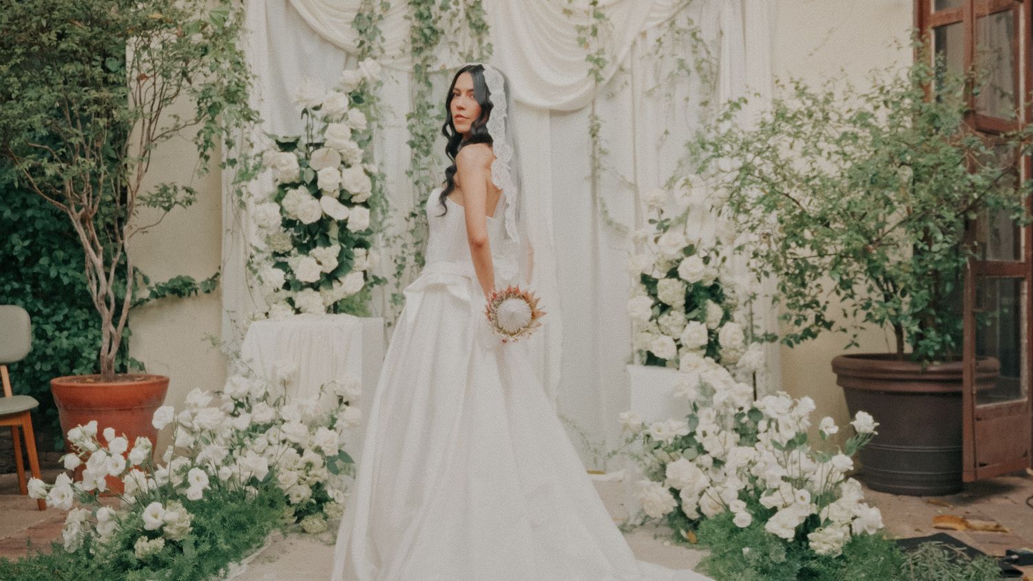 Bride in elegant white ball gown poses among lush white floral arrangements and greenery at a romantic wedding ceremony backdrop.