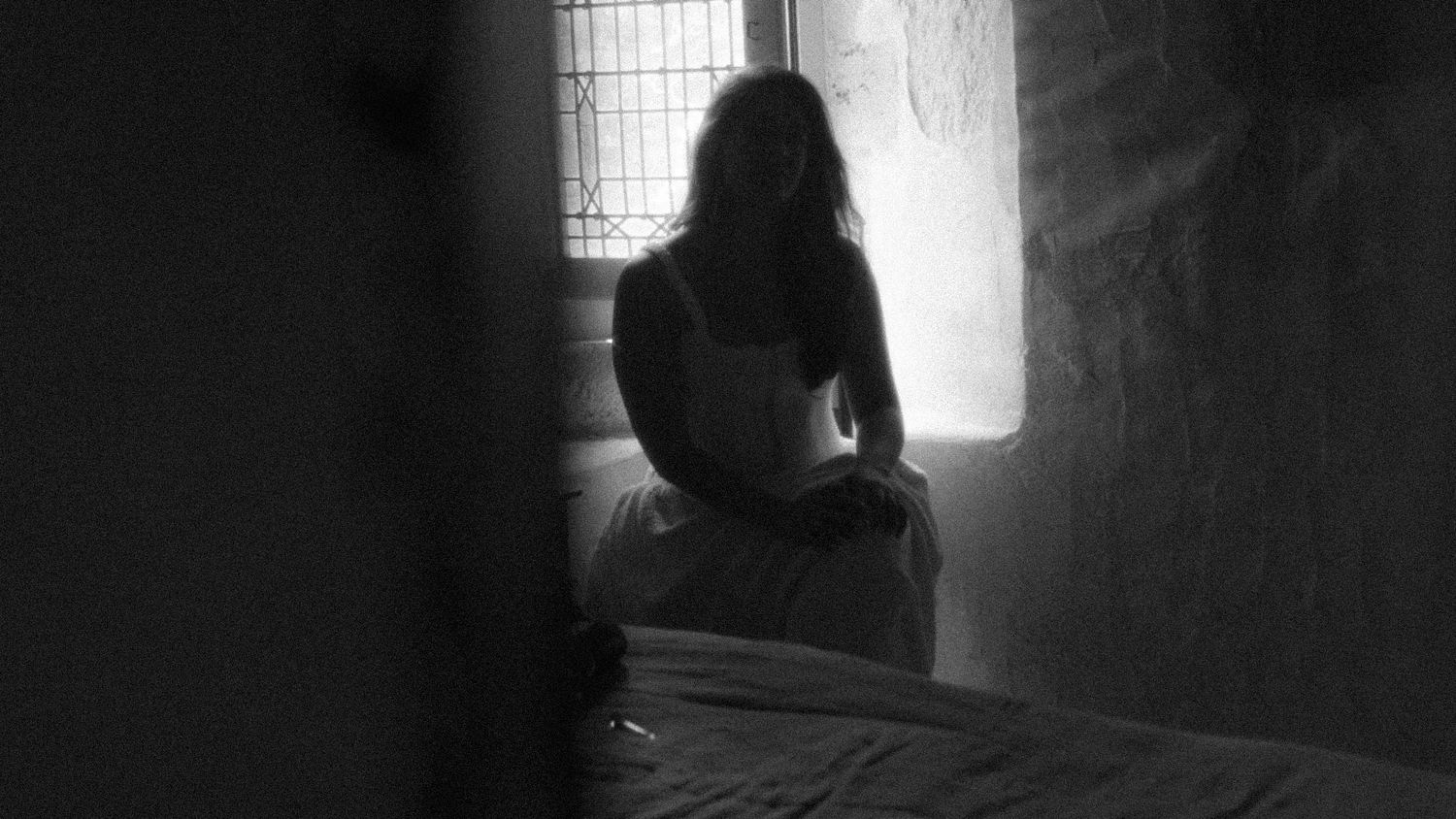 Black and white photo of a woman sitting on a bed near a window in a dimly lit room.