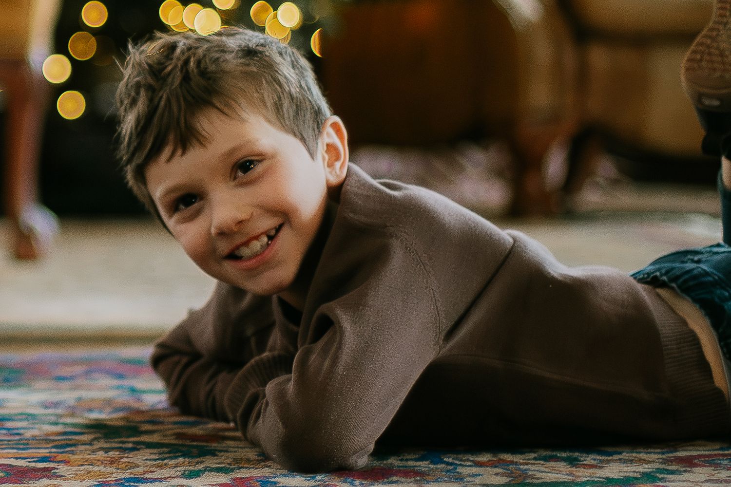 Smiling young boy lying on colorful rug with bokeh Christmas lights in background.