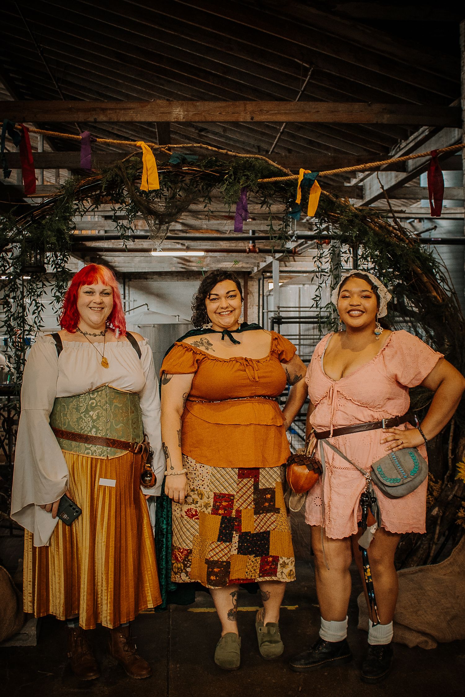 Three women in fantasy costumes pose under a decorated branch archway at the Outer Realms Market event.