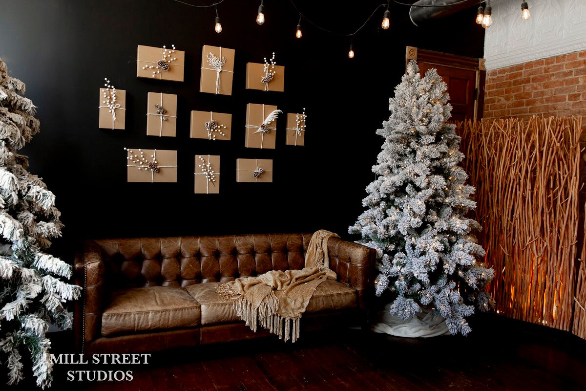 Festive studio with flocked Christmas trees, wrapped gifts on dark wall, and leather sofa with cozy throw blanket.