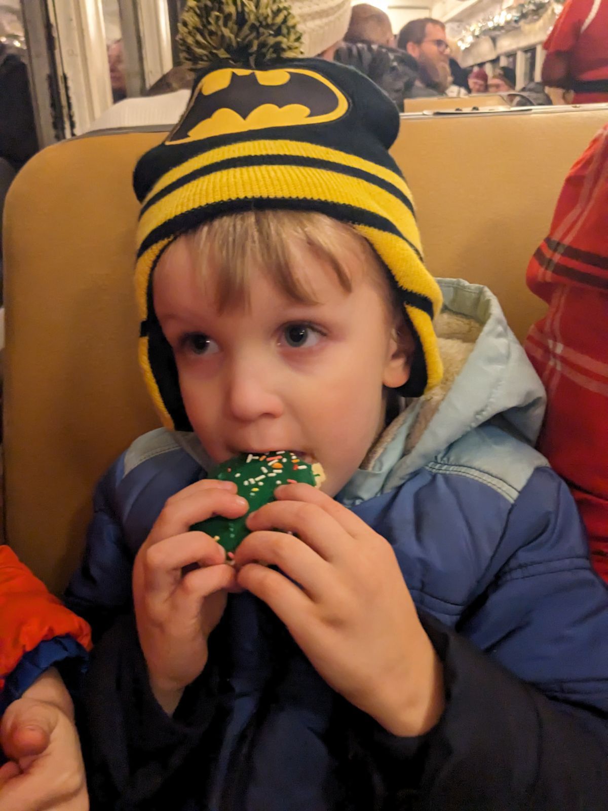 Young boy wearing a Batman winter hat eating a decorated holiday cookie at an indoor event.