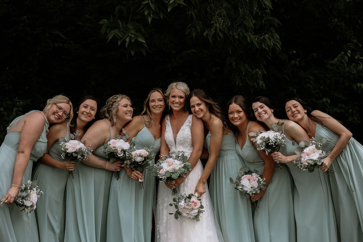 Bride poses with seven bridesmaids in sage green gowns holding white bouquets before lush greenery.