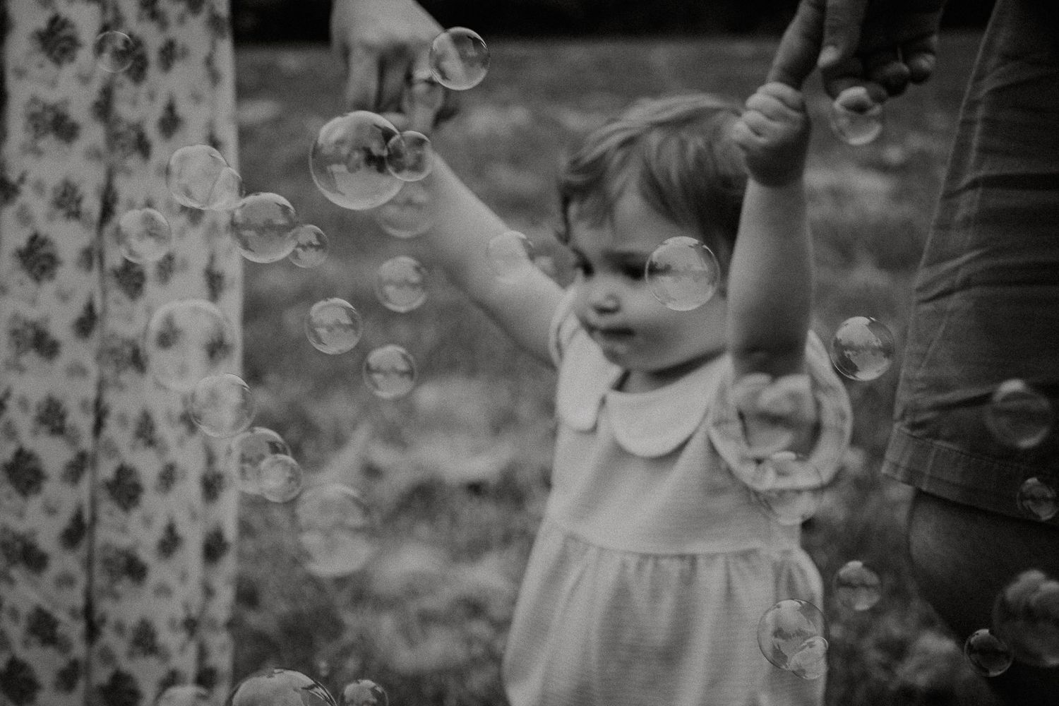 Black and white photo of a toddler girl reaching up to touch floating soap bubbles outdoors.
