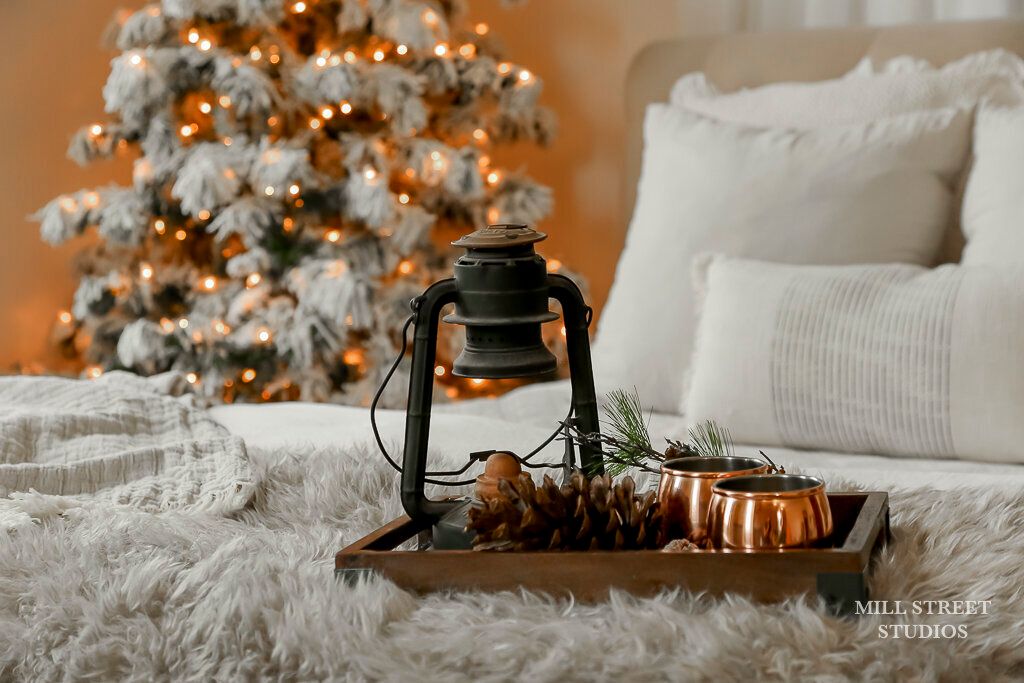 Cozy Christmas bedroom with vintage lantern, pine cones, copper mugs on tray, white fur blanket, and glowing snow-flocked tree.