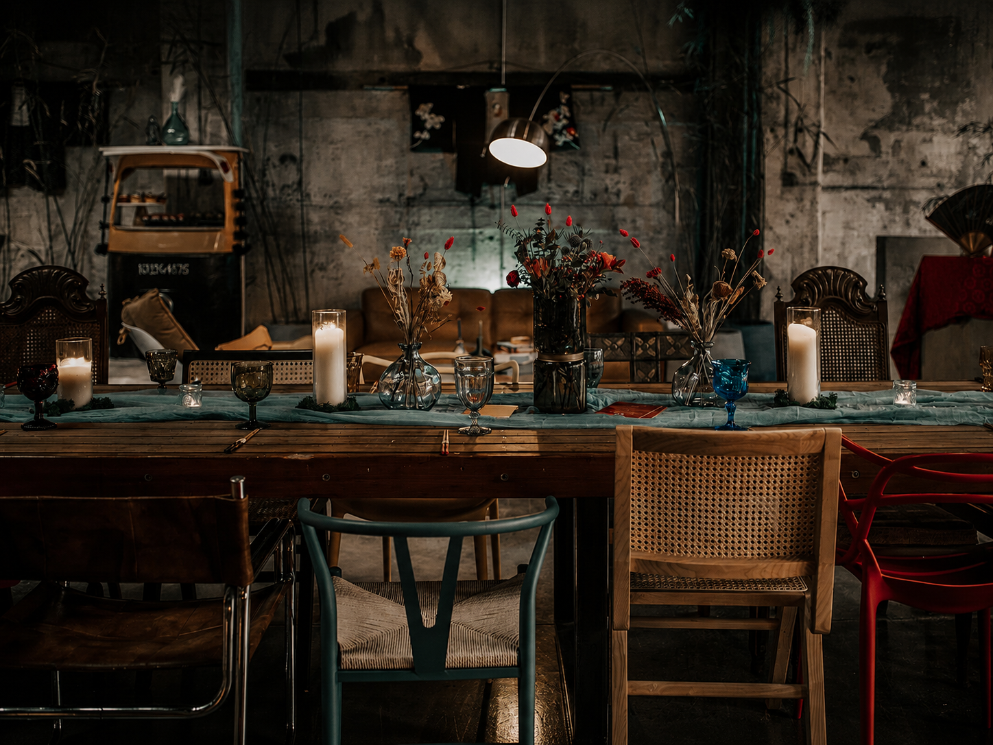 Rustic dining table set with candles and floral decor in a moody, industrial-style space with mismatched vintage chairs.