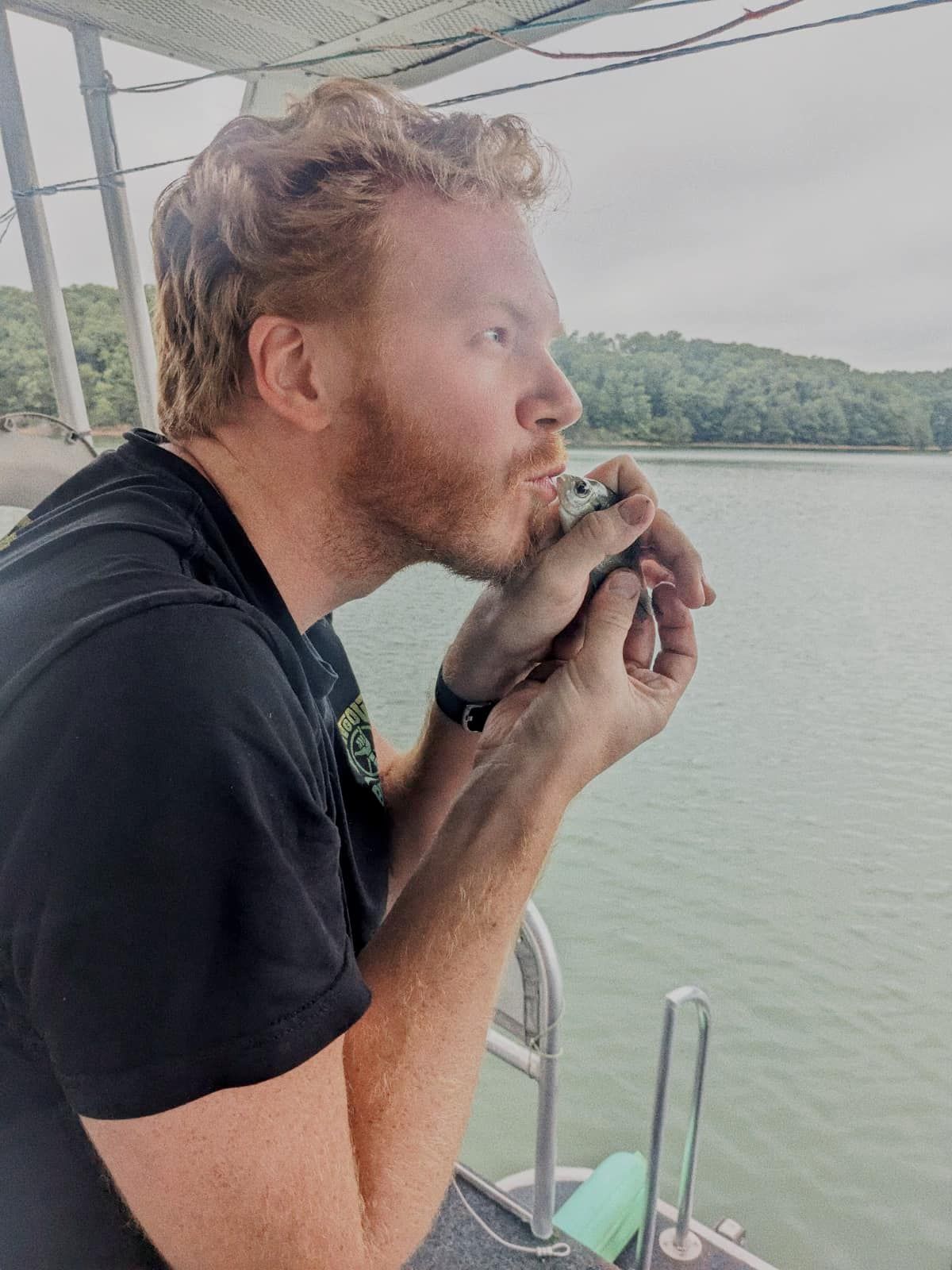 Man smoking on boat deck with scenic lake and trees behind.