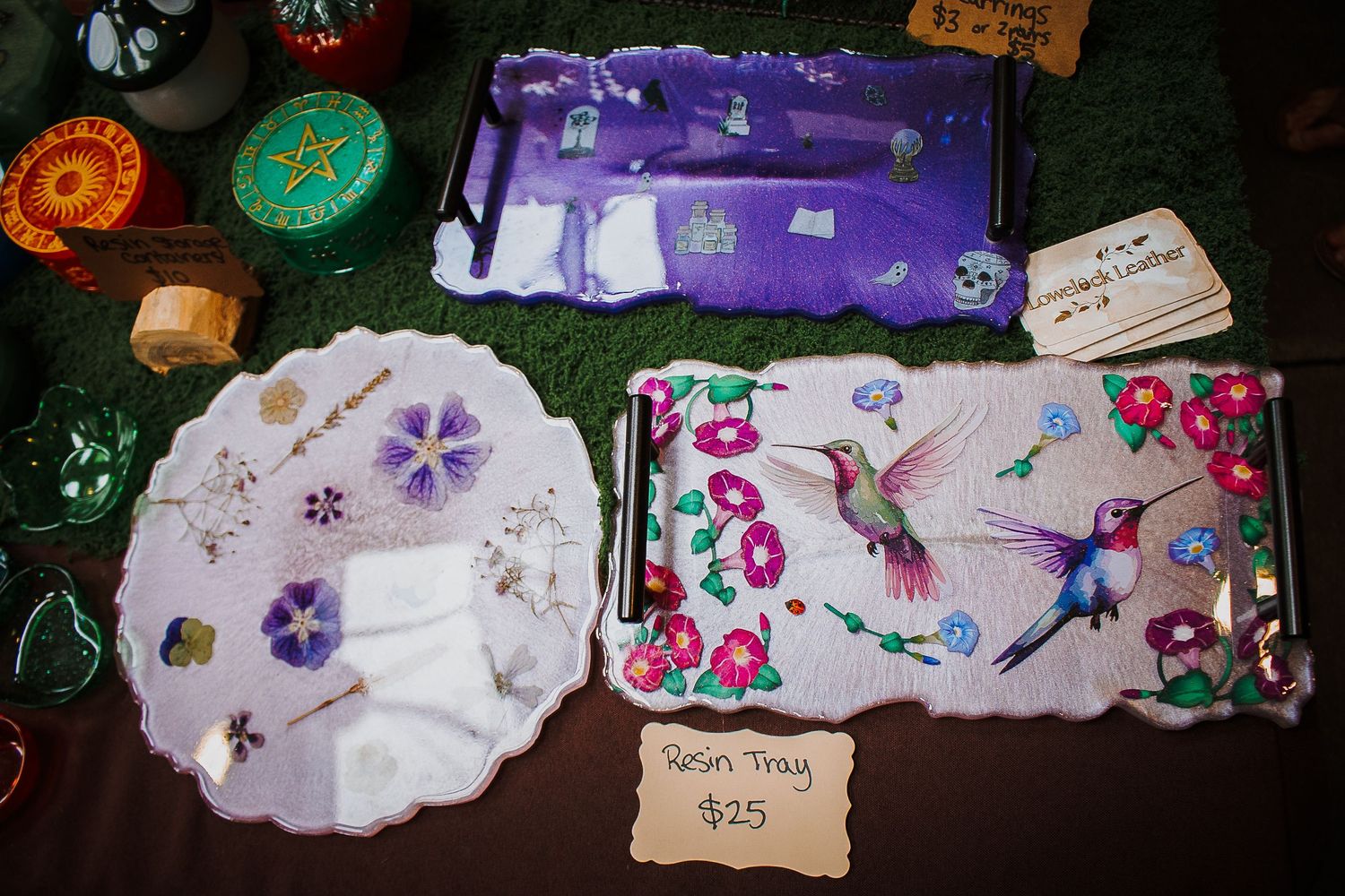 Handmade resin trays priced at $25 featuring floral and hummingbird designs displayed on a green surface.