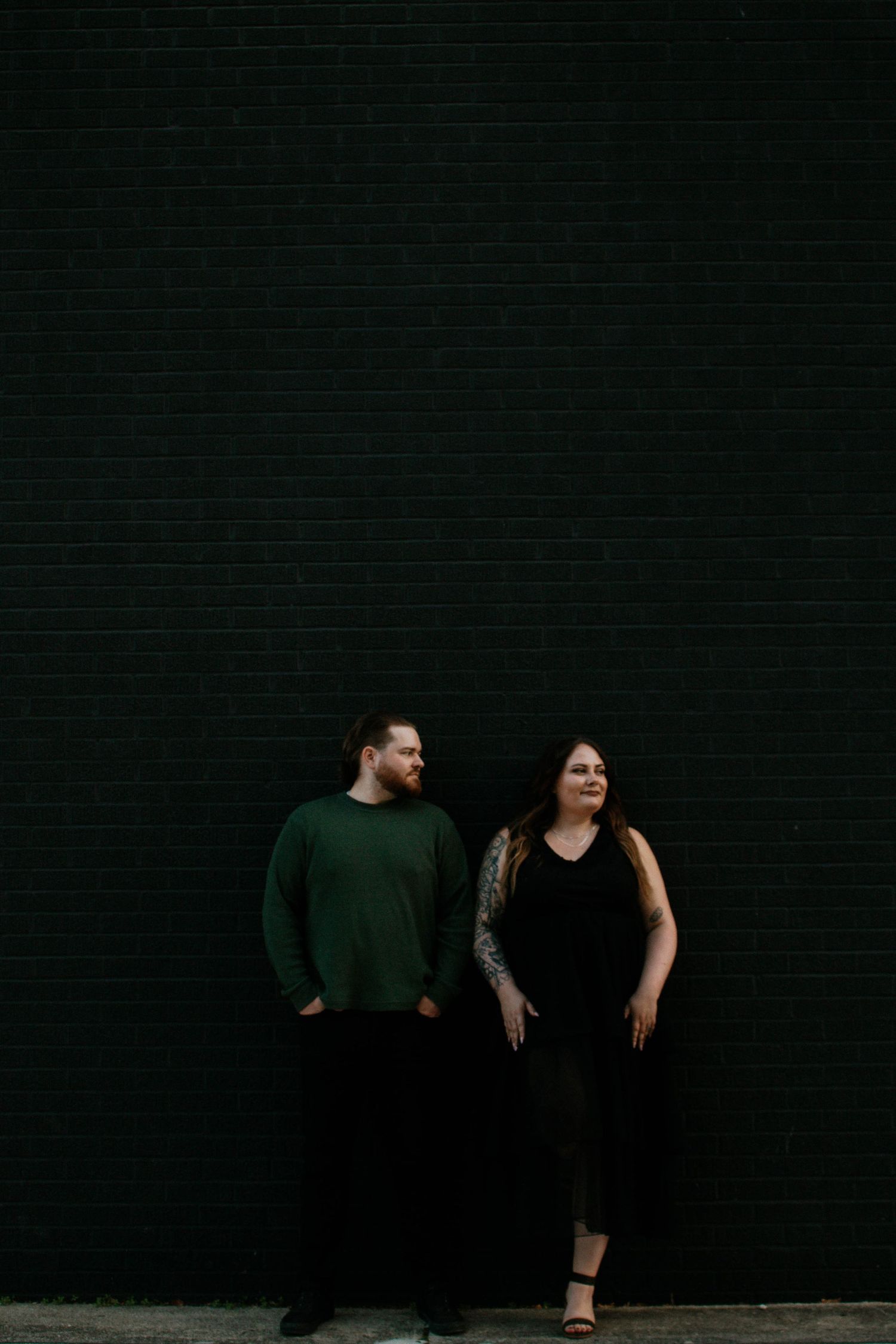 Couple posing against dark green brick wall, man in green sweater, woman in black dress.