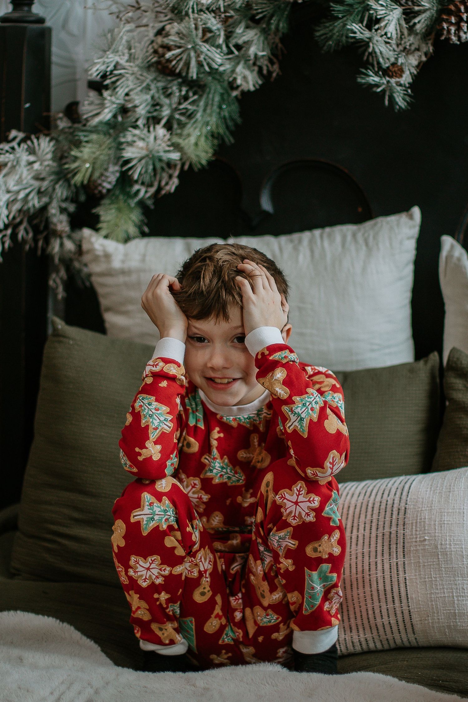 Happy boy in red Christmas pajamas sitting on bed with festive holiday tree decor.