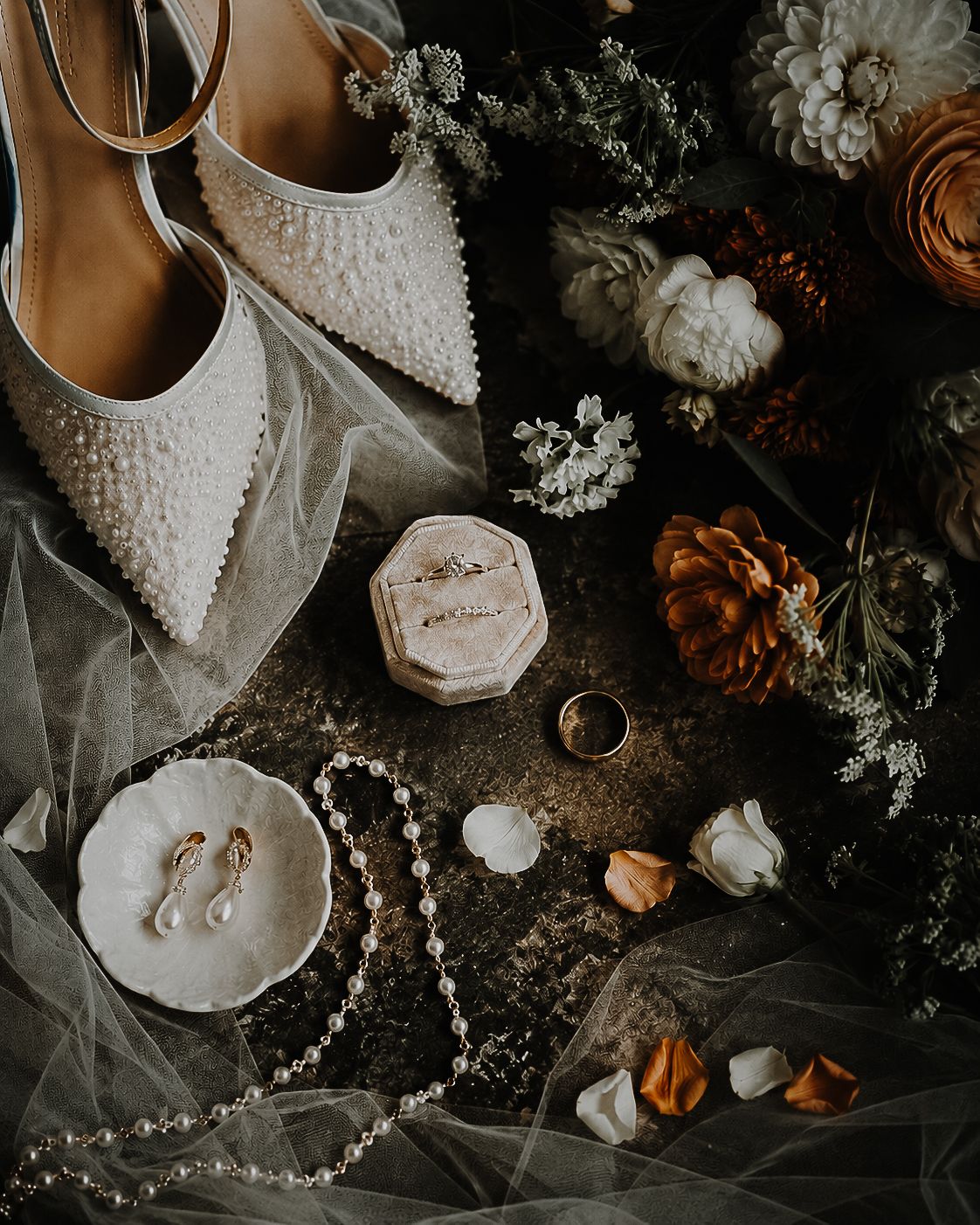 Elegant bridal flat lay with pearl heels, ring box, pearl necklace, earrings, wedding bands, and autumn floral accents.