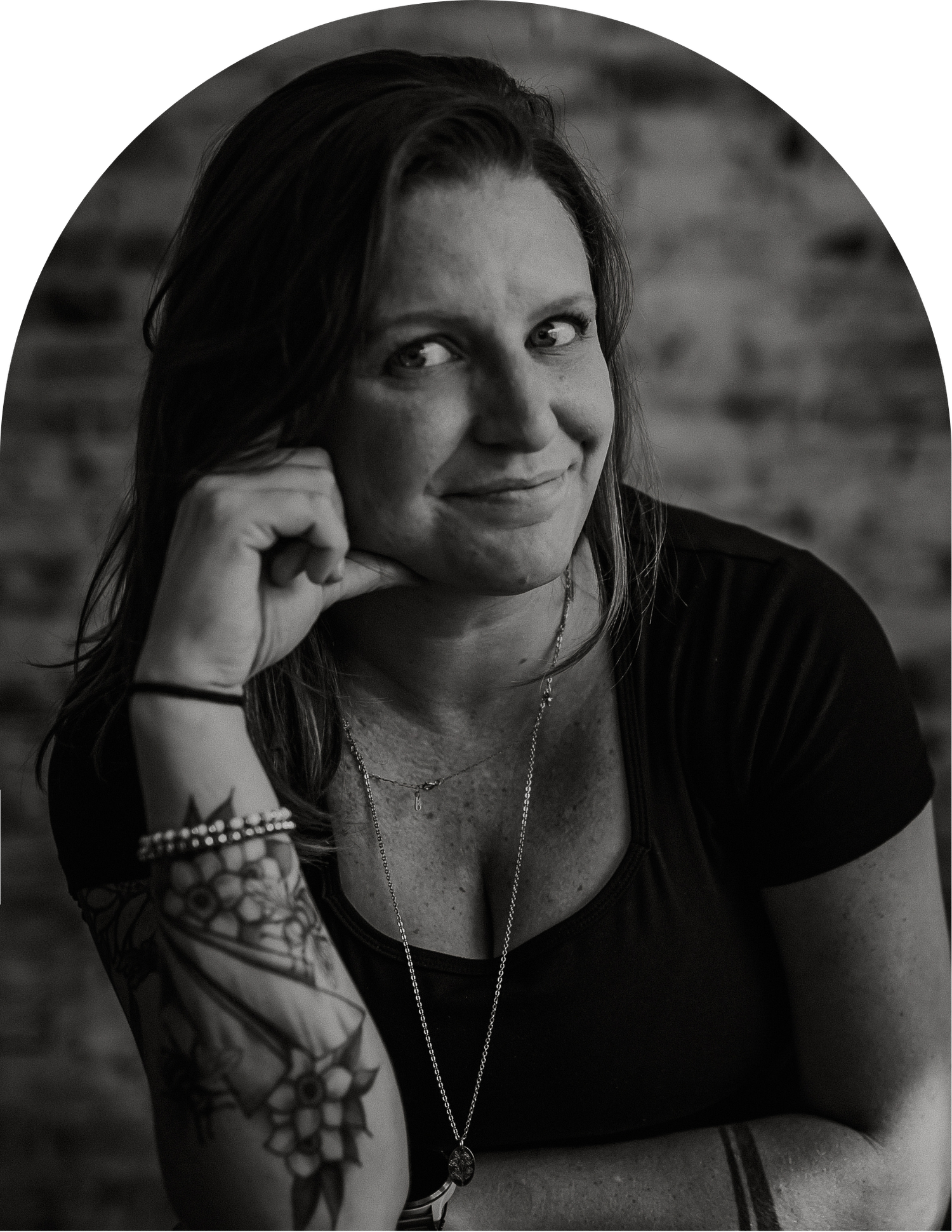 Black and white portrait of a smiling woman with long hair and a floral forearm tattoo, resting her chin on her hand against a brick wall.