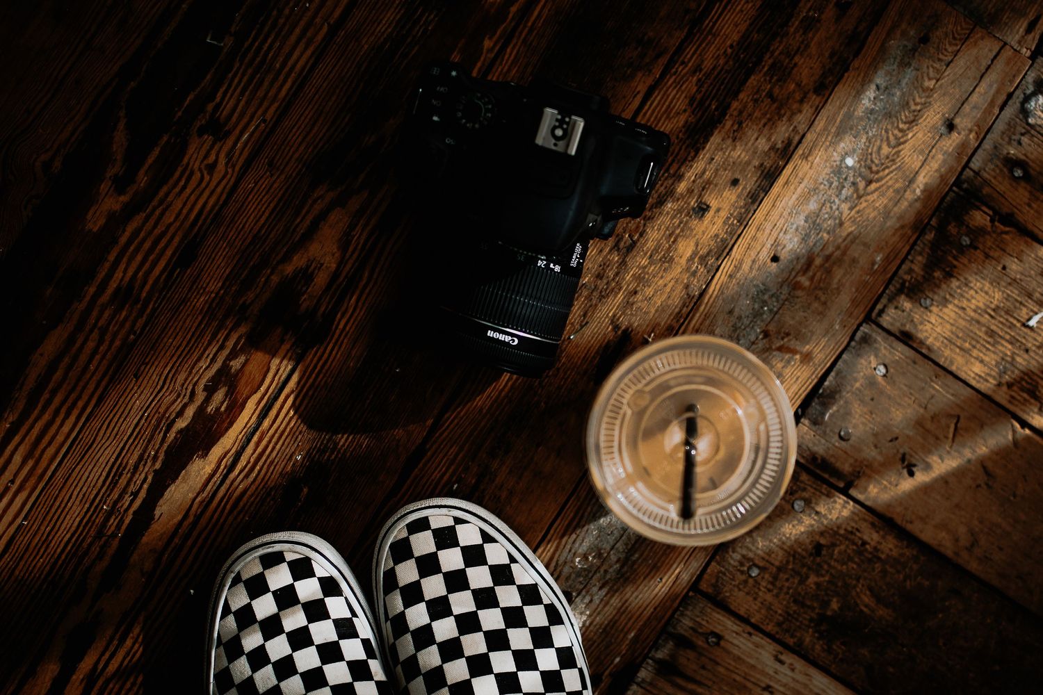 Black and white checkered slip-on sneakers, a camera, and an iced drink with a straw on a dark rustic wooden floor.