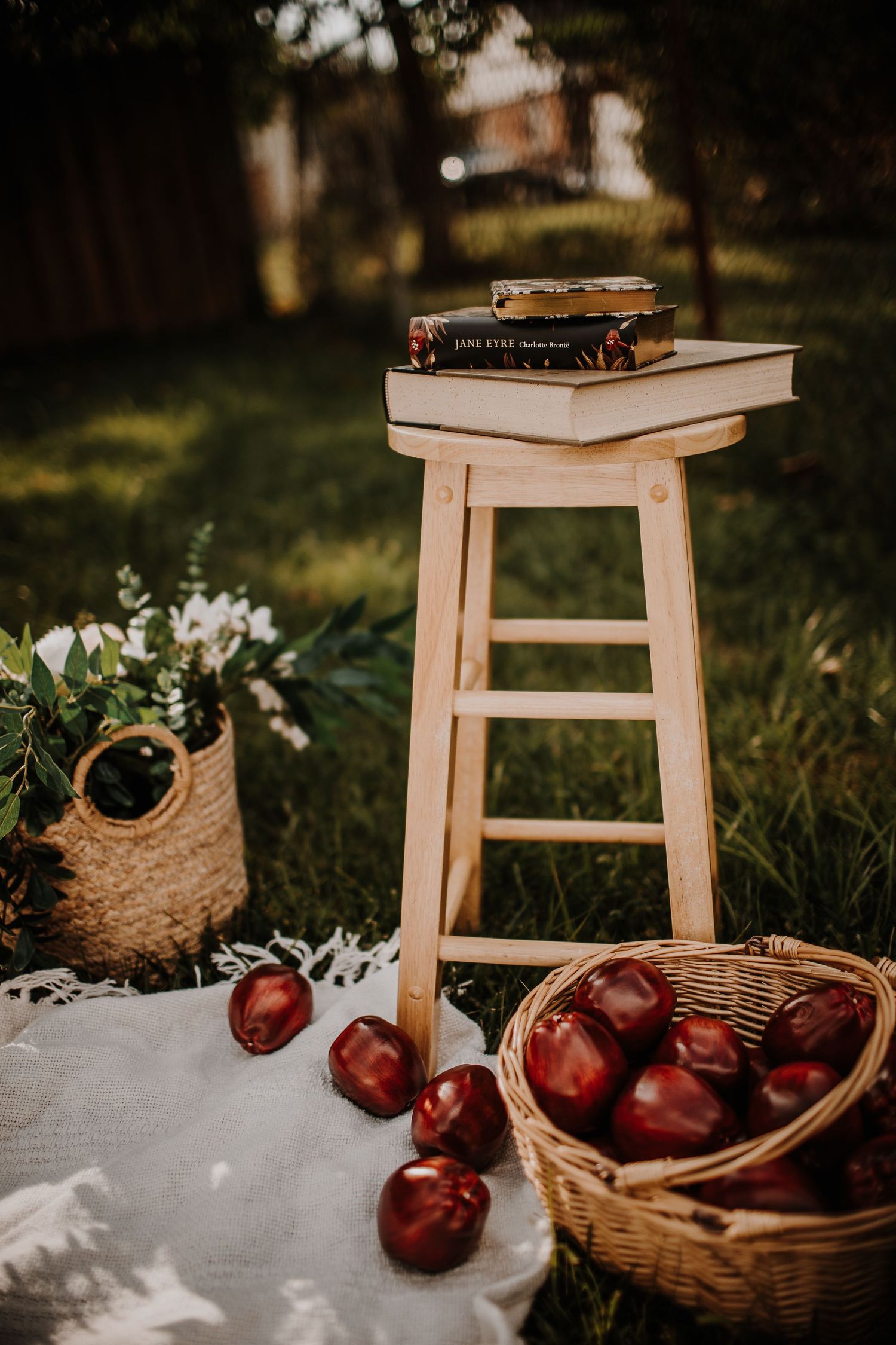 Wooden stool with stacked books including Jane Eyre, red apples in wicker basket, and floral tote bag on white blanket outdoors.