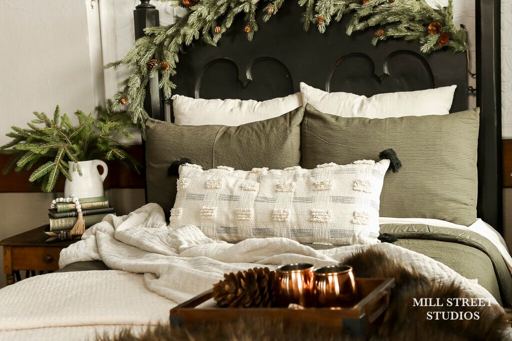 Cozy Christmas bedroom with green pillows, white textured bedding, pine garland headboard, and a wooden tray with candles and pinecones.