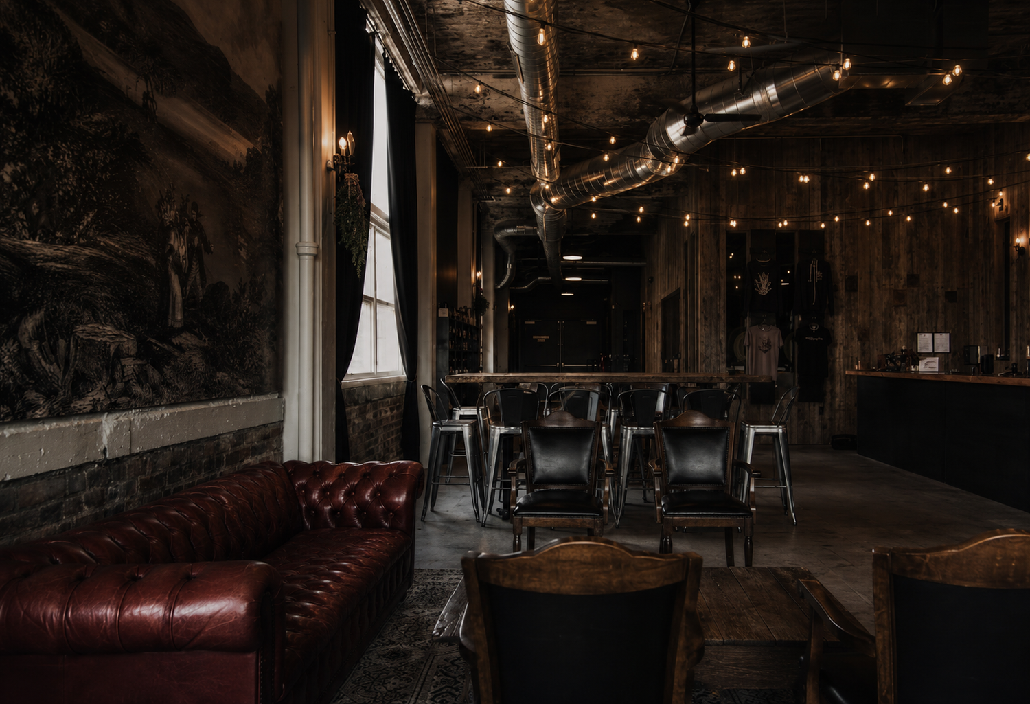 Moody bar interior with red leather Chesterfield sofas, rustic walls, and warm string lights overhead.