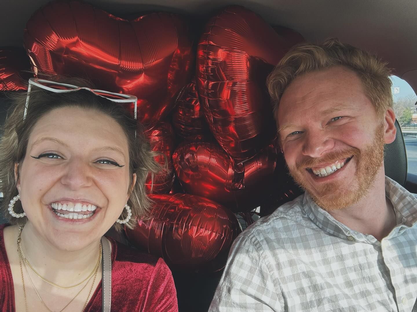 Smiling couple taking selfie in car surrounded by red heart balloons.