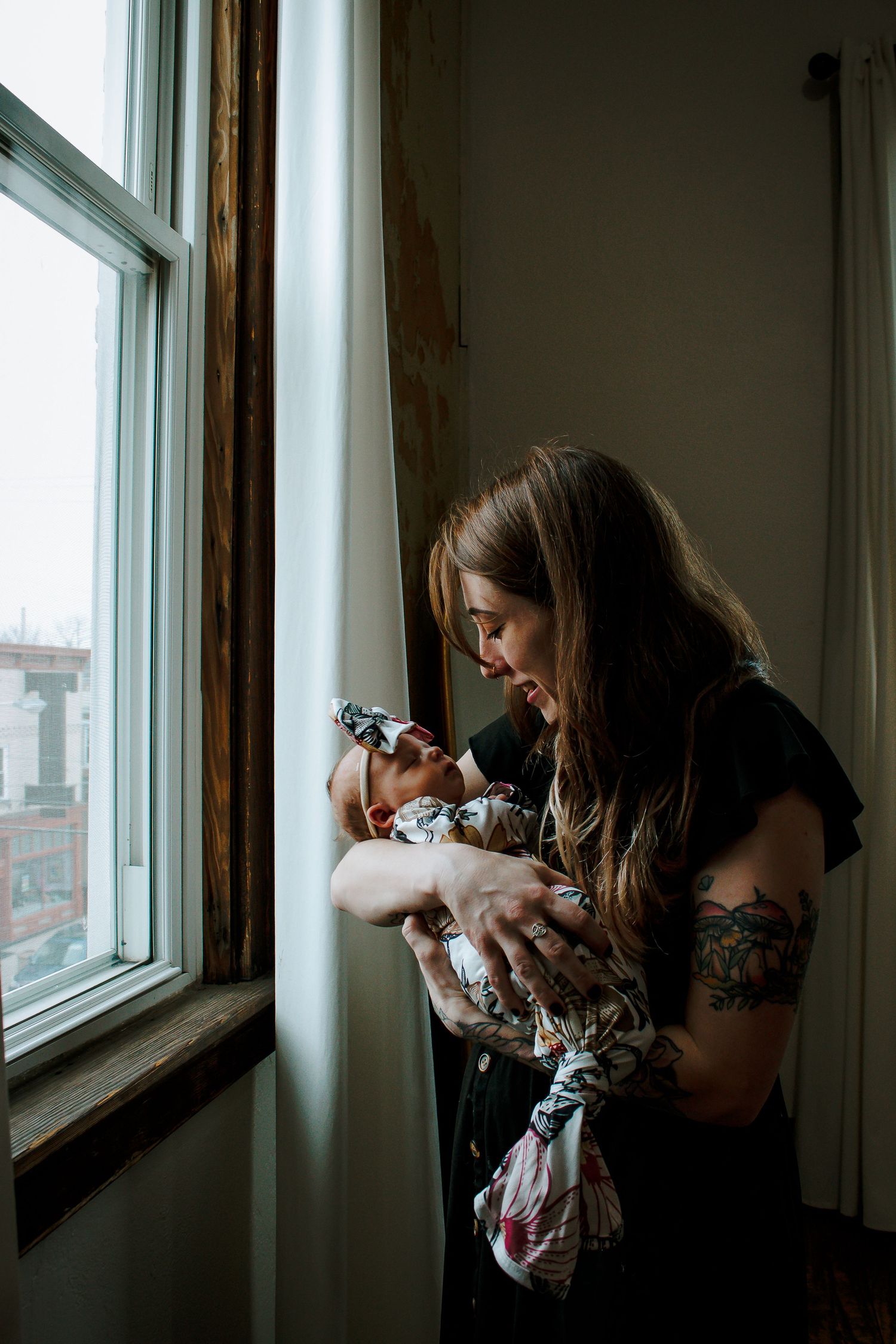 A tattooed mother tenderly holds her newborn baby while standing by a bright window indoors.