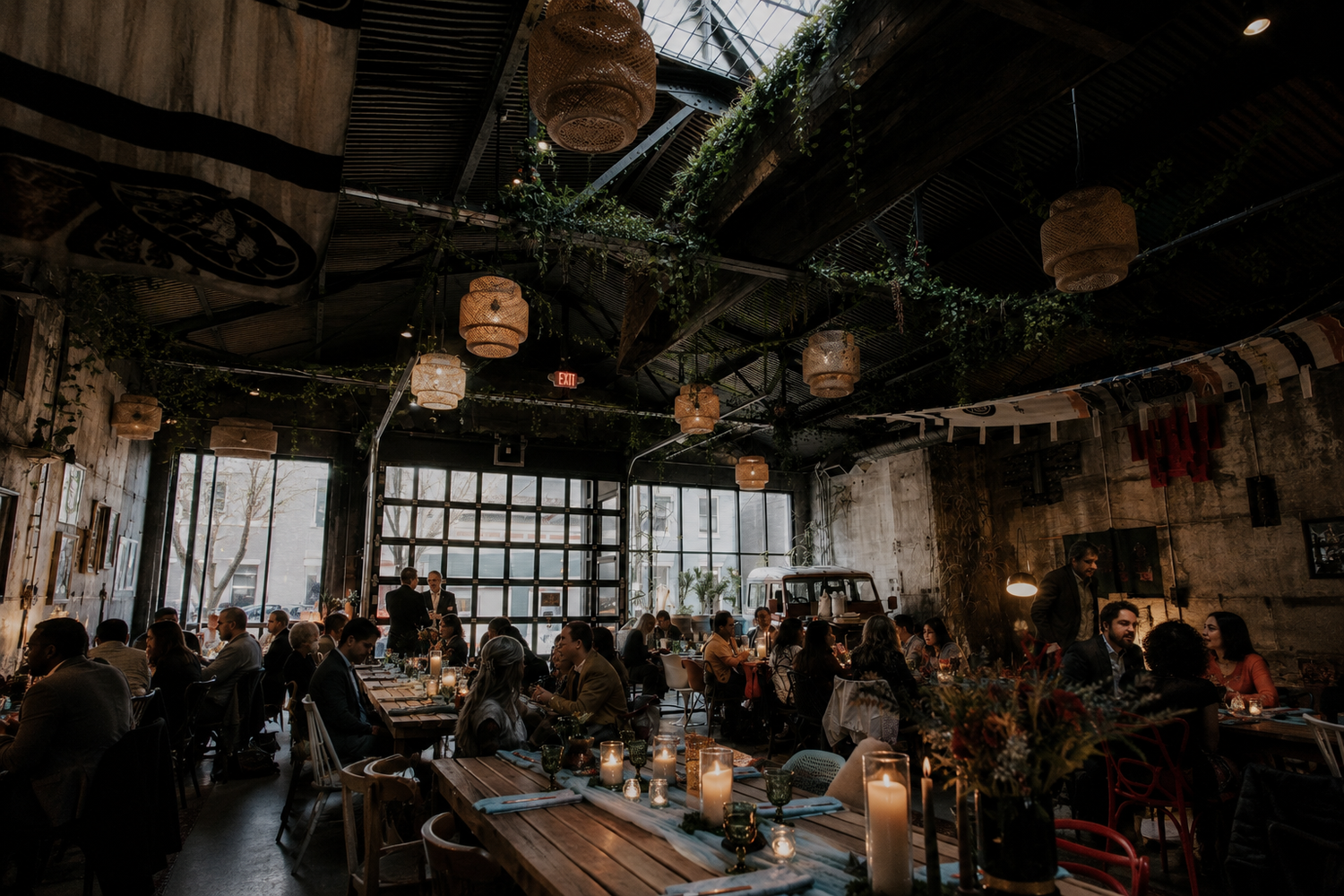 Rustic industrial wedding venue with hanging rattan lights, lush greenery, candlelit tables, and guests dining in a cozy atmosphere.