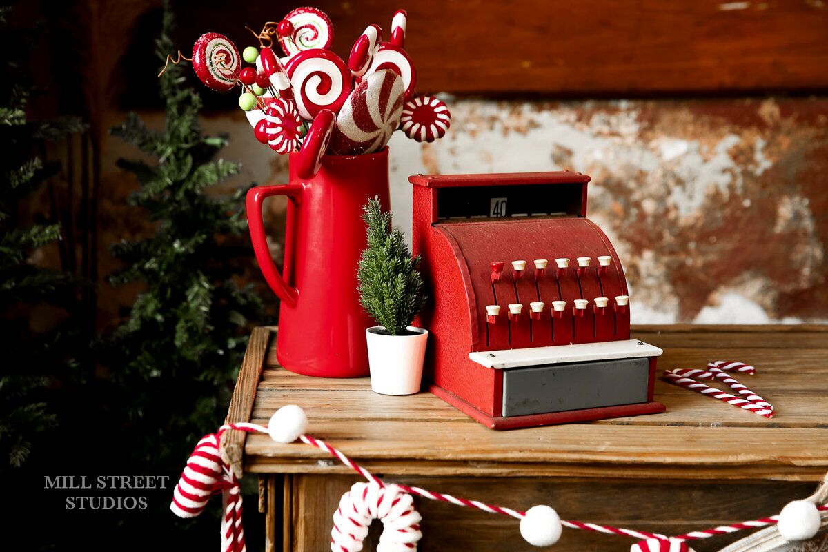 Red vintage cash register with candy canes and peppermint decorations on a rustic wooden table with Christmas greenery.