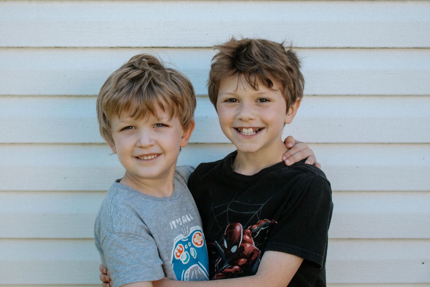 Two young brothers smiling and hugging each other outdoors against a white siding background.