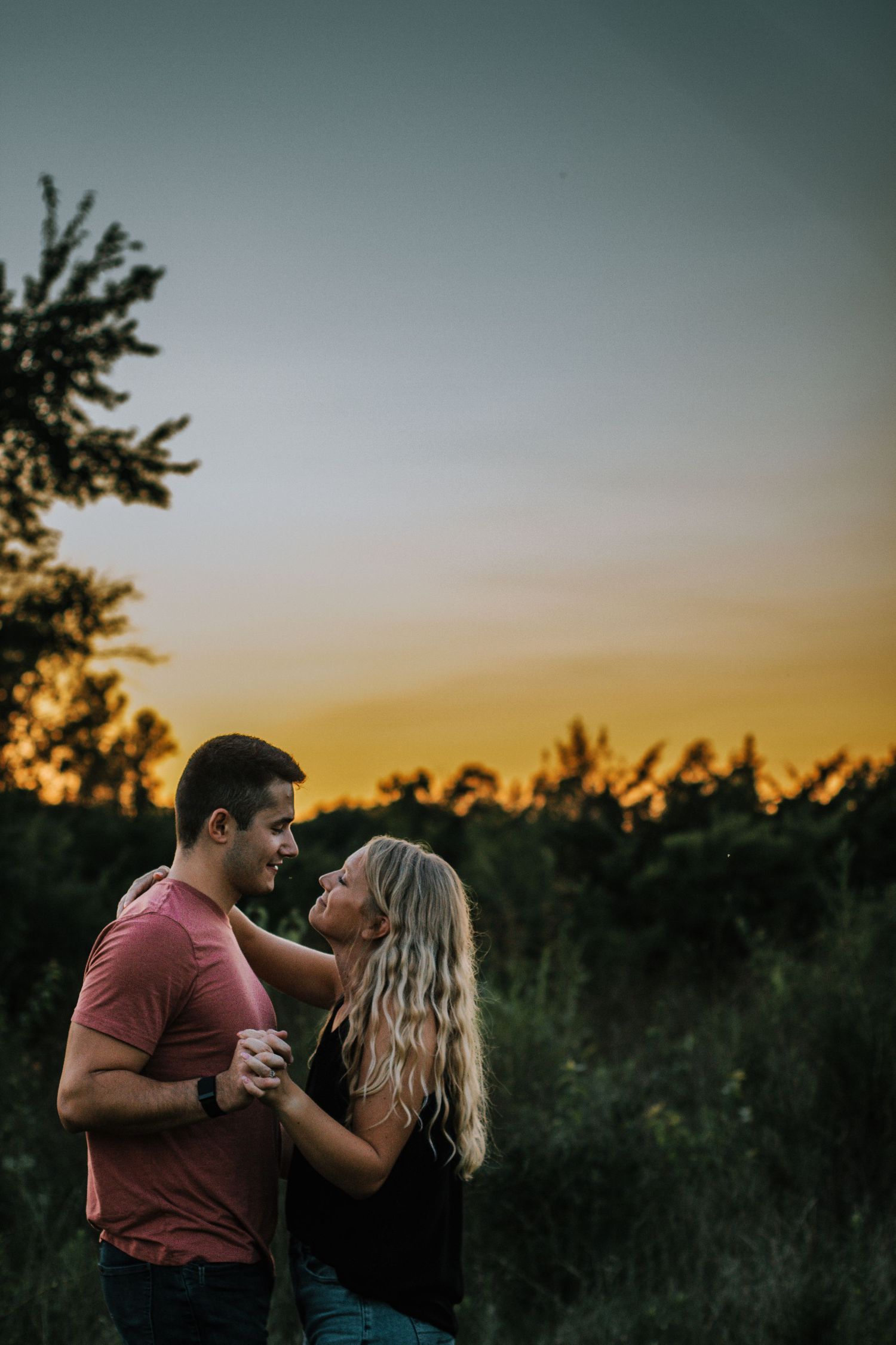 A romantic couple holds hands and gazes at each other against a vibrant sunset sky.
