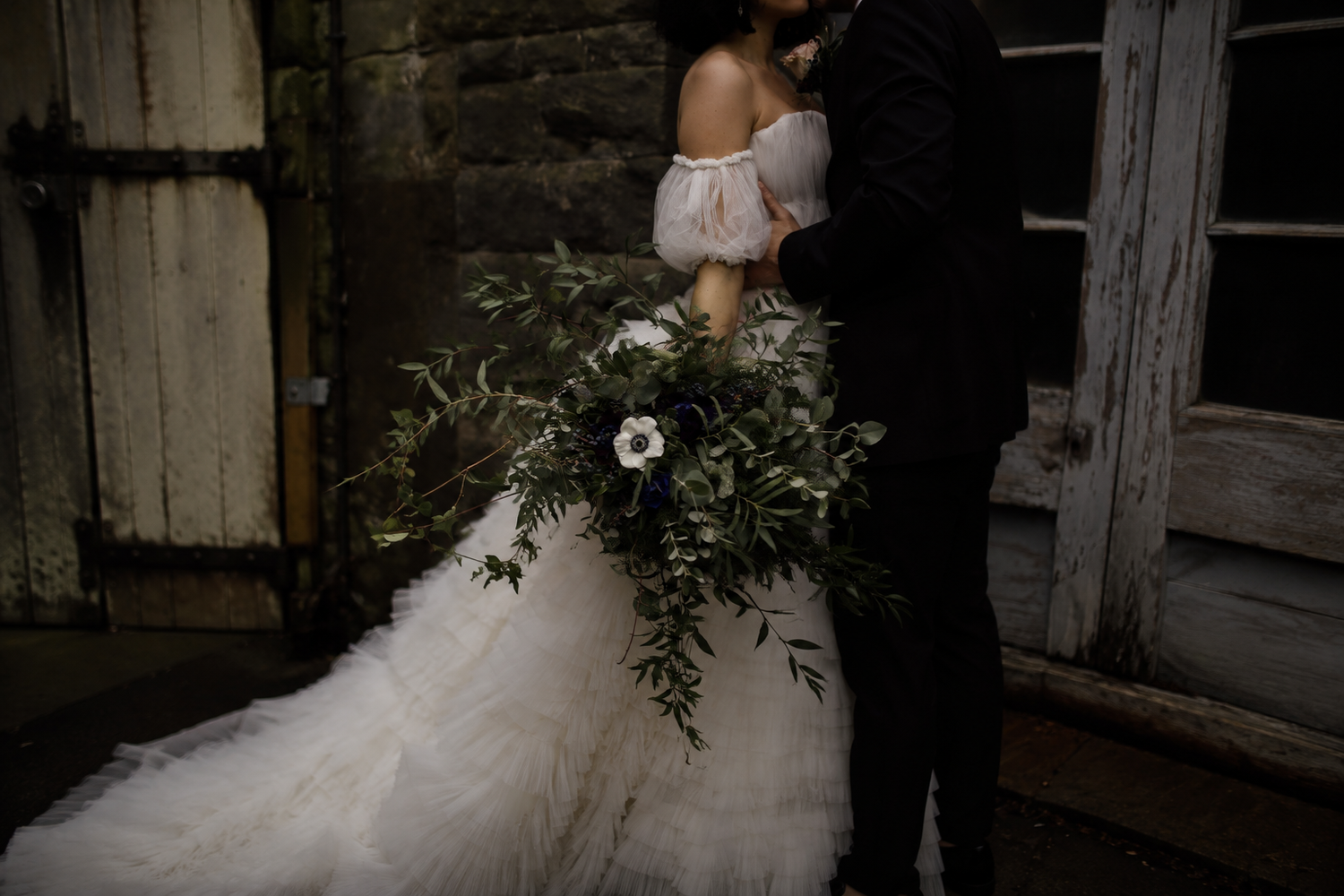 Bride in an off-shoulder tulle gown holding a large greenery bouquet embracing groom in dark suit.