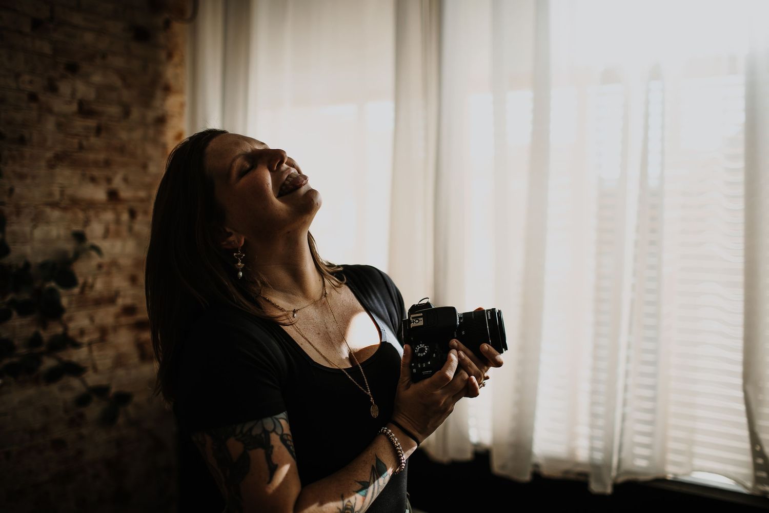 Woman with a tattooed arm laughs with her head tilted back, holding a film camera beside sunlit sheer curtains and a brick wall.