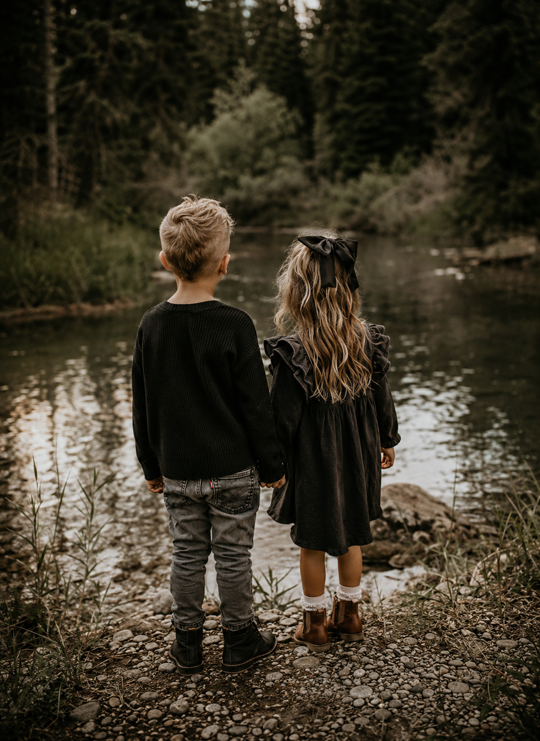 Two young children holding hands by a serene forest river, dressed in matching black outfits, viewed from behind.