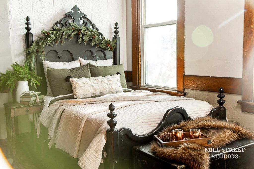 Dark ornate bed with green garland headboard, layered neutral bedding, and cozy holiday decor in a warmly lit bedroom.