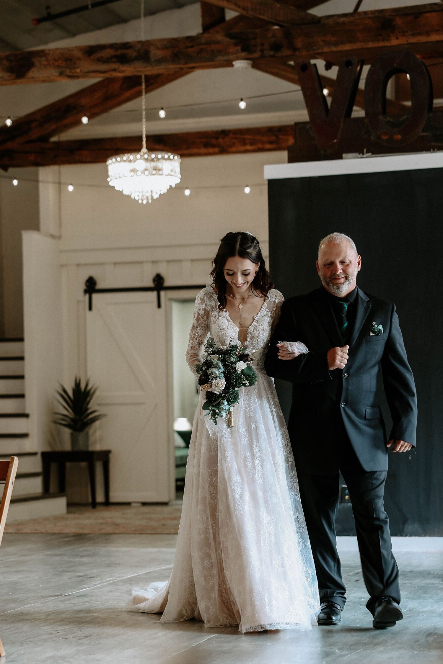 Bride in lace gown holding bouquet walks down aisle with older man in dark suit inside rustic wedding venue.