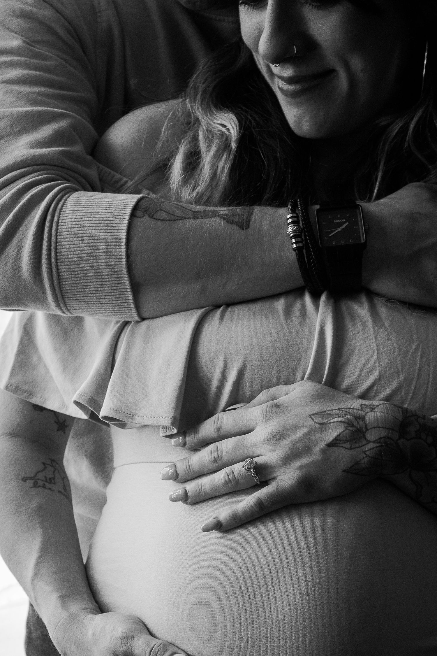 A black and white photo of a couple embracing a pregnant belly, showing tattoos and a watch.