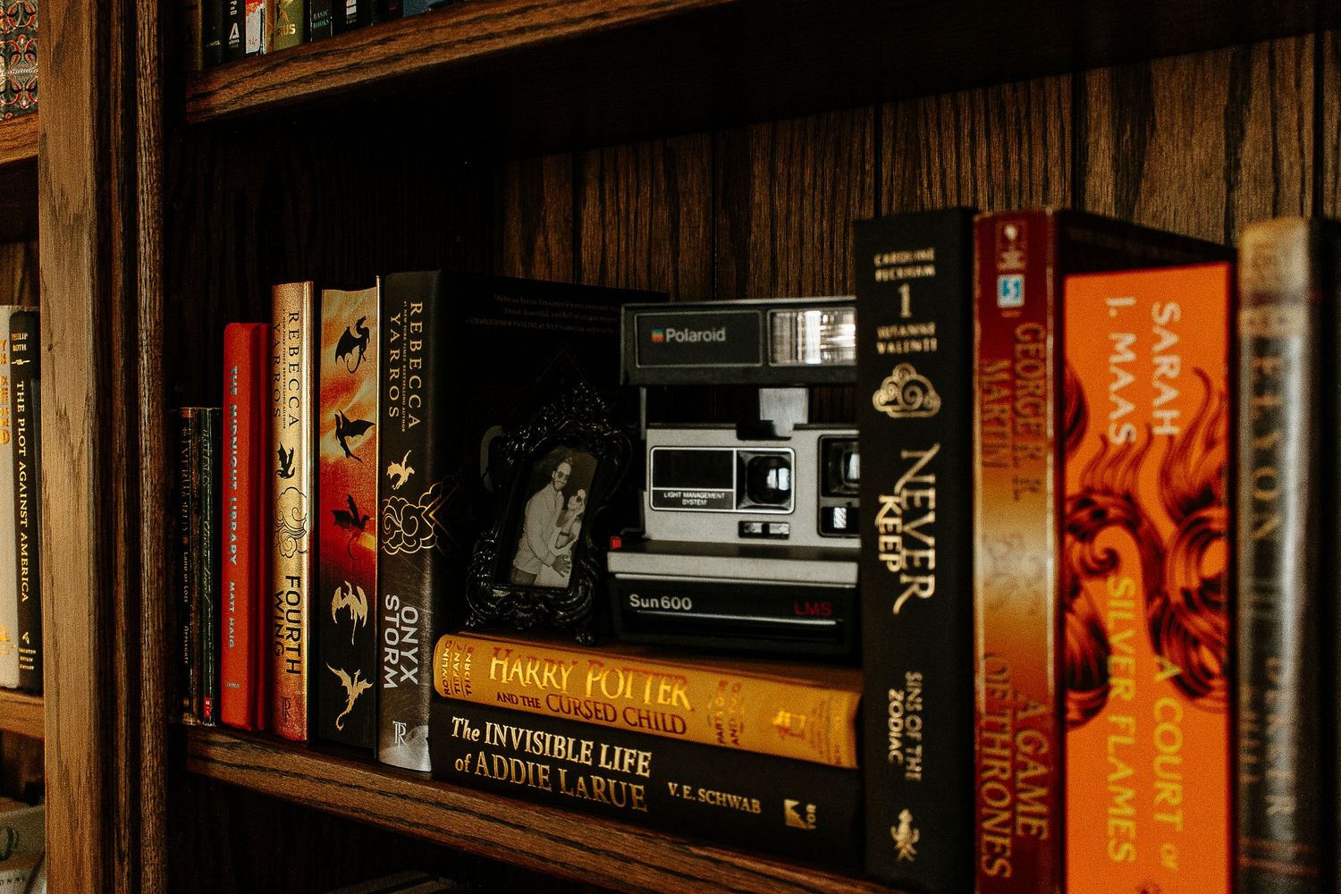 A Polaroid camera sits among colorful books including The Invisible Life of Addie LaRue on a wooden shelf.