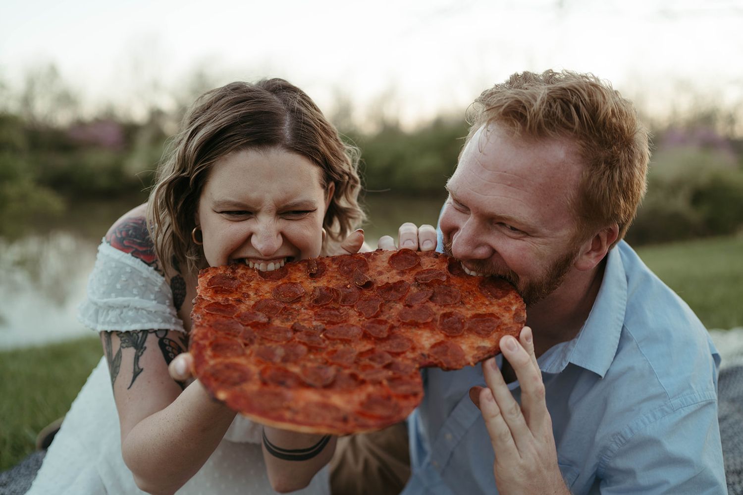 Happy couple laughing while sharing a large pepperoni pizza slice outdoors near a pond.