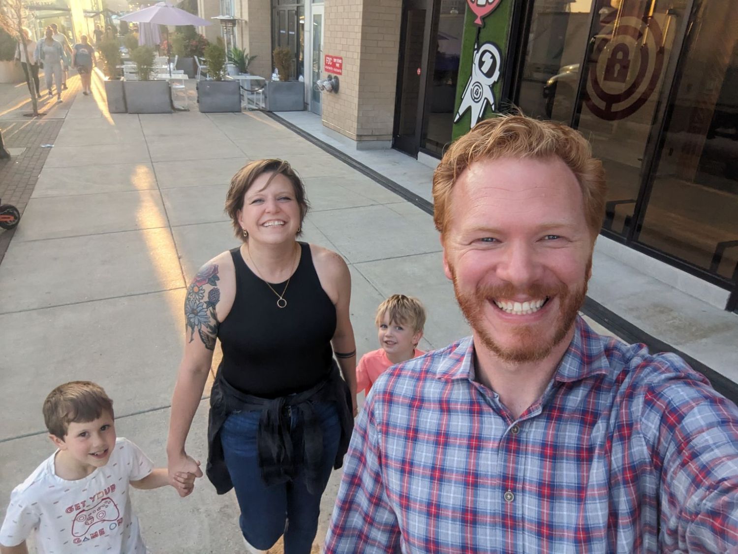 Smiling man takes selfie with family walking on sunny urban sidewalk.